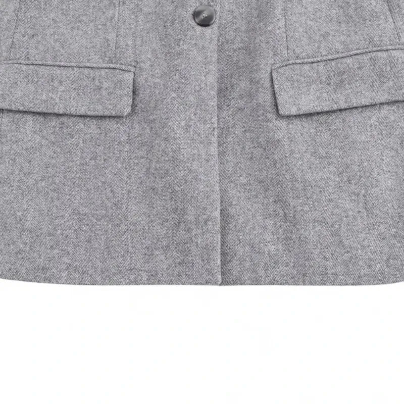 Close-up da parte inferior do blazer feminino cinza, com botão frontal único e bolsos chapados com lapela, exibindo a textura da peça.