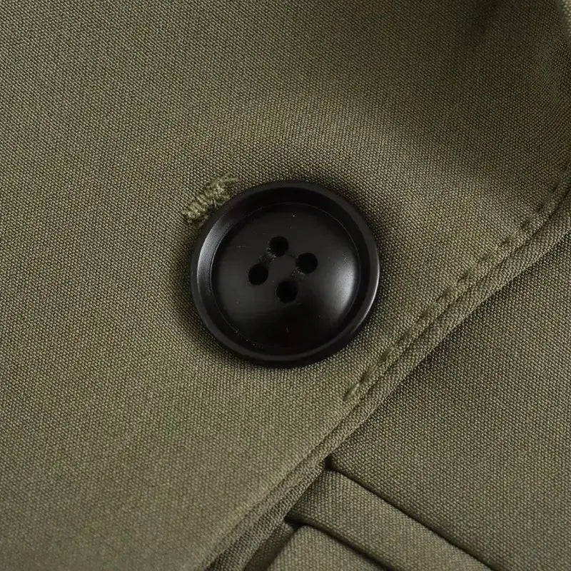 Close-up de botão redondo preto com quatro furos, costurado no tecido verde liso do blazer feminino de alfaiataria verde.