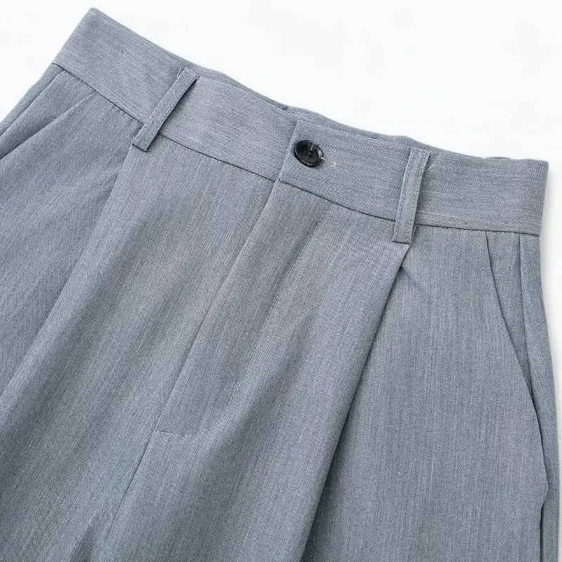 Close-up da cintura alta da calça alfaiataria feminina cinza claro, destacando o botão preto frontal, presilhas de cinto e pregas.