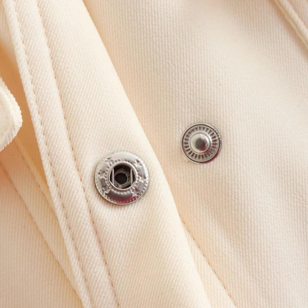Close-up do fechamento da jaqueta bomber feminina off-white, exibindo dois botões de pressão metálicos prateados abertos.