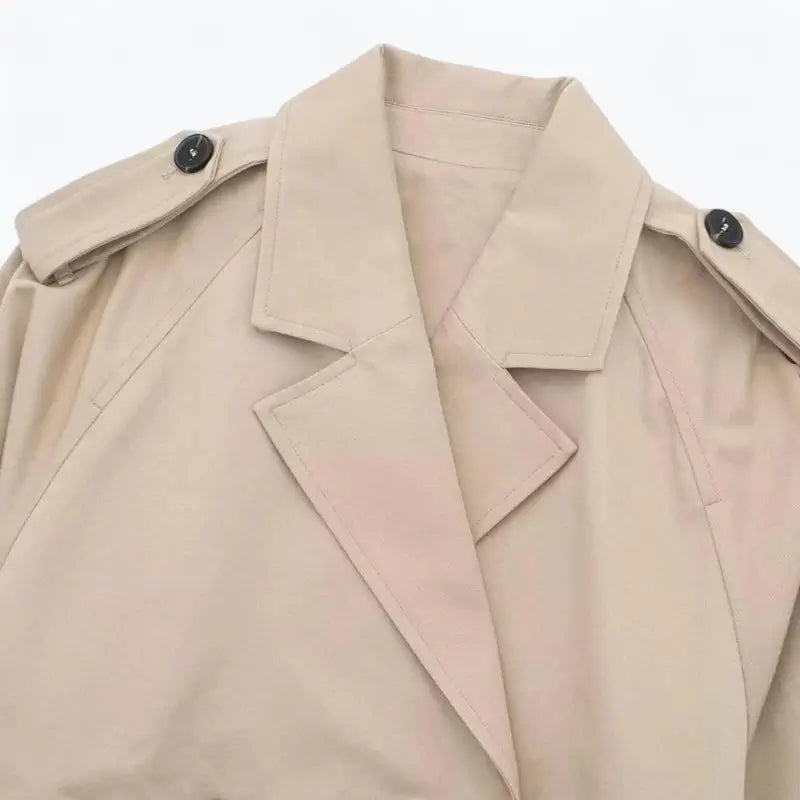 Close-up da gola e lapelas da jaqueta feminina cropped bege estilo trench coat, detalhando as alças dos ombros com botões pretos.