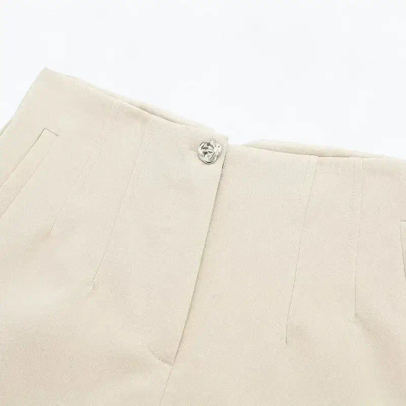 Close-up da cintura de short feminino de alfaiataria em tom bege claro, destacando o botão decorativo no fechamento frontal e a textura lisa do tecido.