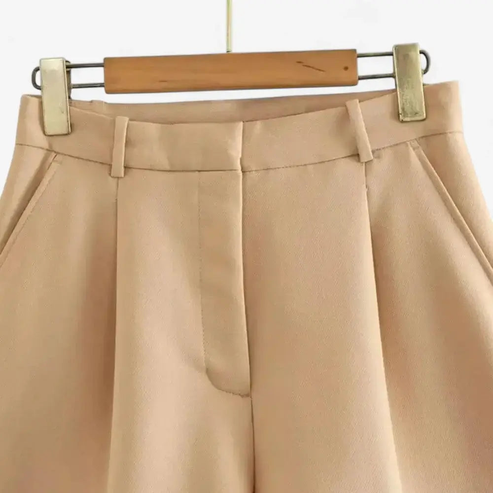 Short alfaiataria feminino bege, modelo cintura alta com pregas frontais bem marcadas e bolsos laterais discretos, pendurado em cabide de madeira.